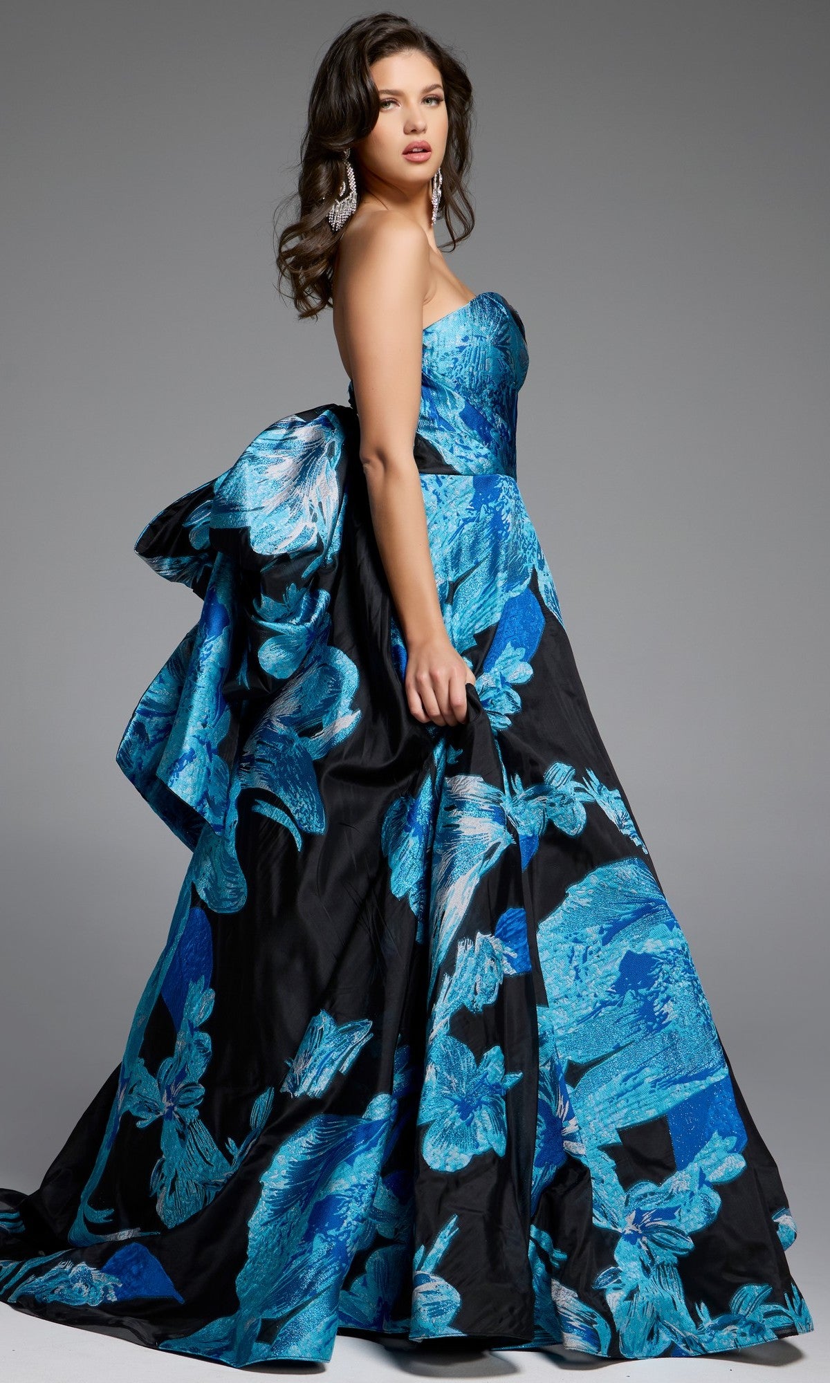 A woman wears the Jovani 42436 Long Prom Dress, a strapless floral ball gown with bold blue patterns and an oversized back bow, standing against a plain gray background.